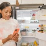 Kobieta przy otwartej lodówce zamawia produkty na telefonie