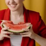 Uśmiechnięta kobieta w czerwonej marynarce trzyma kawałek arbuza na żółtym tle