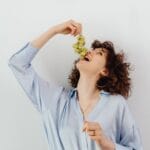 Uśmiechnięta kobieta w niebieskiej bluzce je zielone winogrona w domu