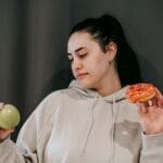 Zamyślona kobieta w bluzie wybiera między zielonym jabłkiem a donutem